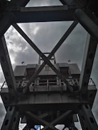 A large, industrial steel structure with a complex lattice design, viewed from below against a cloudy sky. The metal framework includes crossbeams and riveted joints, suggesting the architecture of a bridge or crane.