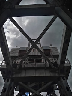 A large, industrial steel structure with a complex lattice design, viewed from below against a cloudy sky. The metal framework includes crossbeams and riveted joints, suggesting the architecture of a bridge or crane.