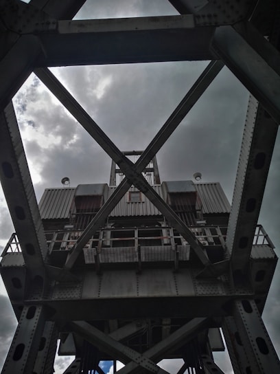 A large, industrial steel structure with a complex lattice design, viewed from below against a cloudy sky. The metal framework includes crossbeams and riveted joints, suggesting the architecture of a bridge or crane.