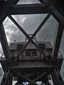 A large, industrial steel structure with a complex lattice design, viewed from below against a cloudy sky. The metal framework includes crossbeams and riveted joints, suggesting the architecture of a bridge or crane.