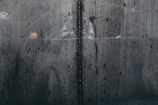A close-up view of a textured, industrial metal surface with visible welding marks running vertically. The metal displays various stains and discolorations, giving it an aged and worn appearance. Light reflections and subtle rust patches add to the surface's character.