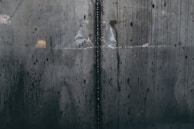 A close-up view of a textured, industrial metal surface with visible welding marks running vertically. The metal displays various stains and discolorations, giving it an aged and worn appearance. Light reflections and subtle rust patches add to the surface's character.