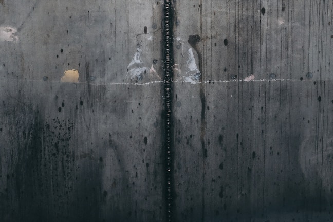 A close-up view of a textured, industrial metal surface with visible welding marks running vertically. The metal displays various stains and discolorations, giving it an aged and worn appearance. Light reflections and subtle rust patches add to the surface's character.