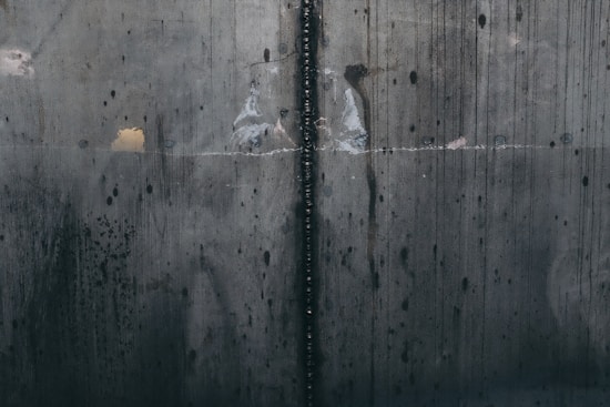 A close-up view of a textured, industrial metal surface with visible welding marks running vertically. The metal displays various stains and discolorations, giving it an aged and worn appearance. Light reflections and subtle rust patches add to the surface's character.