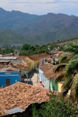 A picturesque view of a vibrant town nestled against a backdrop of lush green mountains. The foreground features a series of colorful colonial-style buildings with tiled roofs, set along a narrow, cobblestone street. Palm trees and greenery add to the tropical ambiance.