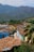 A picturesque view of a vibrant town nestled against a backdrop of lush green mountains. The foreground features a series of colorful colonial-style buildings with tiled roofs, set along a narrow, cobblestone street. Palm trees and greenery add to the tropical ambiance.