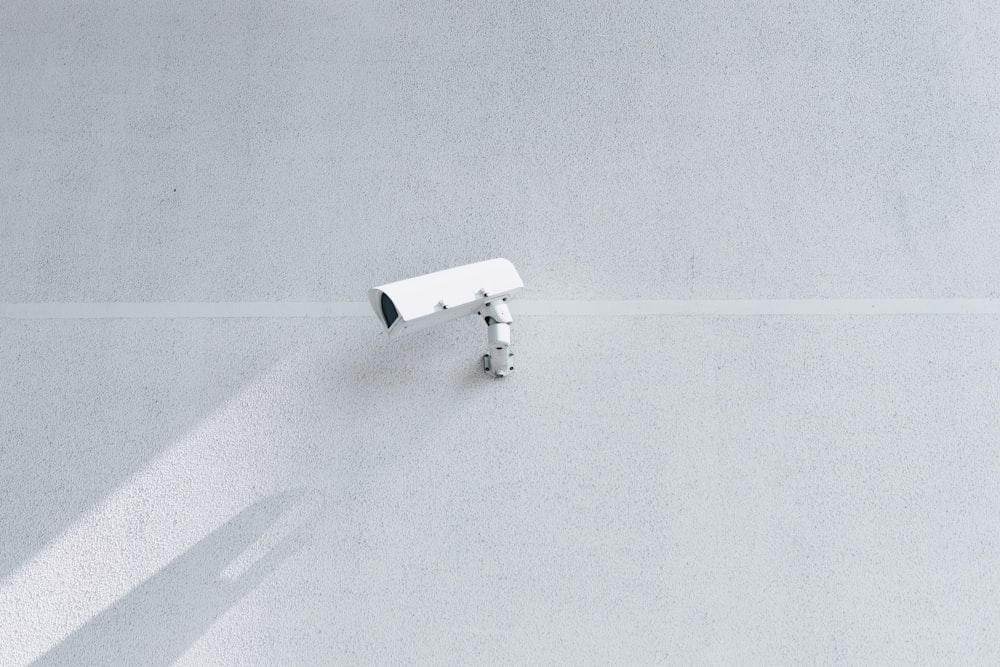 A white surveillance camera casts a shadow on a blank white wall