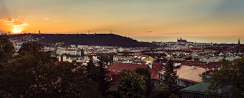 A picturesque cityscape at sunset, featuring a blend of traditional red-roofed buildings and lush greenery in the foreground. The skyline is dominated by historical architecture, including a grand cathedral silhouetted against a vibrant sky. The sun sets dramatically on the horizon, casting a warm glow over the entire scene.