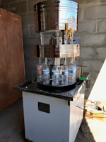 A large industrial bottle capping machine is in operation. Several plastic water bottles are positioned on a rotating platform beneath the metallic capping mechanism. The setup is located in a room with bare concrete block walls, and there is a wooden board leaning against the wall. Sunlight casts shadows on the floor, and there are various tools and materials scattered around, including a cardboard box with cables.