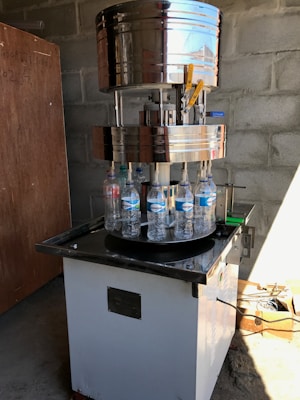 A large industrial bottle capping machine is in operation. Several plastic water bottles are positioned on a rotating platform beneath the metallic capping mechanism. The setup is located in a room with bare concrete block walls, and there is a wooden board leaning against the wall. Sunlight casts shadows on the floor, and there are various tools and materials scattered around, including a cardboard box with cables.