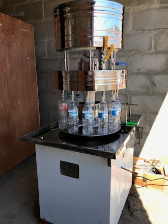 A large industrial bottle capping machine is in operation. Several plastic water bottles are positioned on a rotating platform beneath the metallic capping mechanism. The setup is located in a room with bare concrete block walls, and there is a wooden board leaning against the wall. Sunlight casts shadows on the floor, and there are various tools and materials scattered around, including a cardboard box with cables.