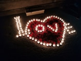 An arrangement of small candles forms the word 'love' surrounded by heart petals on a textured surface. A box is placed near the arrangement, and the scene is dimly lit, creating a warm and intimate atmosphere.
