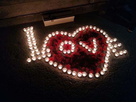 An arrangement of small candles forms the word 'love' surrounded by heart petals on a textured surface. A box is placed near the arrangement, and the scene is dimly lit, creating a warm and intimate atmosphere.