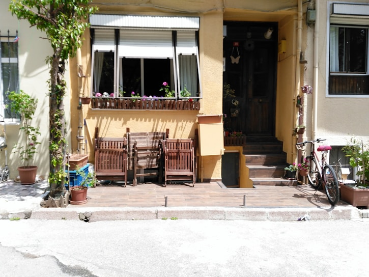 A cozy front porch area with a wooden set of chairs and a small table aligned against a yellow wall. There are potted plants with lush green leaves and some blooming purple flowers. A bicycle is parked on the right side, and a small set of steps leads up to a dark wooden door. The window sill above the chairs is decorated with potted flowers.