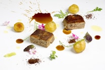 An elegant food presentation featuring two pieces of cooked meat, accompanied by small, round potato croquettes. Herb garnishes and edible flowers are artfully scattered around the plate, with drops and splatters of rich brown sauce adding a creative touch.