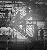 Silhouettes of workers are visible through a mesh screen as they navigate metal scaffolding. The scene is monochromatic, highlighting the intricate network of beams and platforms. The workers, wearing helmets, appear engaged in construction activities, and their forms are partially obscured by the mesh, creating a layered effect.