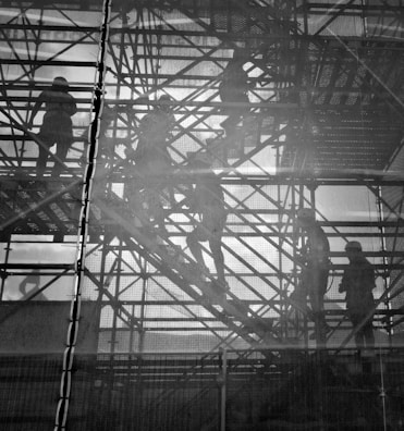 Silhouettes of workers are visible through a mesh screen as they navigate metal scaffolding. The scene is monochromatic, highlighting the intricate network of beams and platforms. The workers, wearing helmets, appear engaged in construction activities, and their forms are partially obscured by the mesh, creating a layered effect.