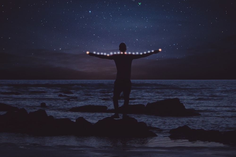 Silhouette einer Person, die auf einem Felsen am Ufer steht und nachts eine Lichterkette spannt