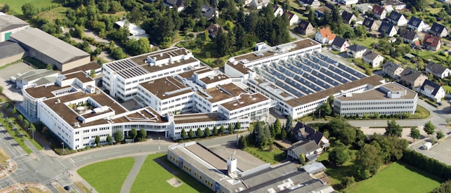 An aerial view of a large, modern industrial or institutional complex with multiple buildings featuring flat, brown roofs and white walls. The complex is surrounded by greenery and other smaller residential structures. There are roads and parking areas around the buildings.