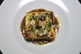 A gourmet dish featuring a layered pasta with melted cheese and herbs, garnished with microgreens and nuts. The dish is presented on a white plate, surrounded by a rich, brown sauce.