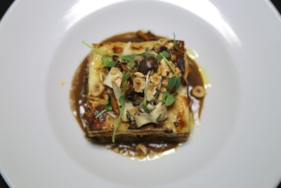 A gourmet dish featuring a layered pasta with melted cheese and herbs, garnished with microgreens and nuts. The dish is presented on a white plate, surrounded by a rich, brown sauce.