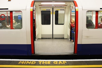 A subway train with its doors open is stationary at a platform. The words 'MIND THE GAP' are painted in yellow on the platform edge. Inside the train, several passengers are visible, including a woman seated by the door holding onto a pole. The interior features red and blue colors with gray flooring.