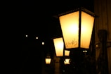 A series of vintage-style street lamps with glowing yellow light along a dark street. Each lamp is housed in a rectangular glass case with a metal frame, mounted on an ornate metal pole. The background is mostly dark with little detail visible.