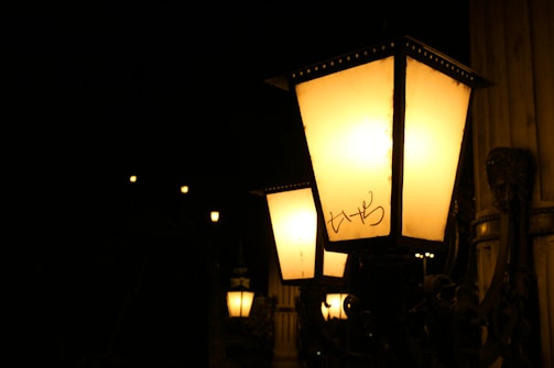 A series of vintage-style street lamps with glowing yellow light along a dark street. Each lamp is housed in a rectangular glass case with a metal frame, mounted on an ornate metal pole. The background is mostly dark with little detail visible.