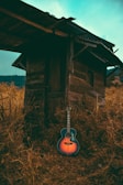Close-up of a rustic guitar resting against a wooden fence with golden wheat in the background.