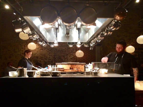 A professional kitchen with two chefs preparing food under a vent hood with hanging pots and pans. The setting is dimly lit, creating a focused and intimate atmosphere. The chefs are working attentively, surrounded by various kitchen utensils and ingredients.