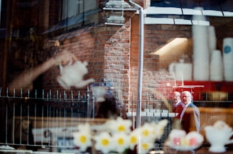 A complex reflection on a glass window shows a brick building with architectural details. In the foreground, there are blurred and reflective images of flowers, a teapot, and a person, possibly a man, walking on a street. The scene is a juxtaposition of indoor and outdoor elements with a surreal overlay.