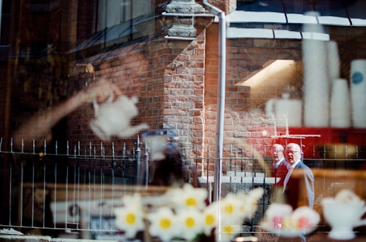 A complex reflection on a glass window shows a brick building with architectural details. In the foreground, there are blurred and reflective images of flowers, a teapot, and a person, possibly a man, walking on a street. The scene is a juxtaposition of indoor and outdoor elements with a surreal overlay.