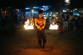 A shirtless man performs a fire-eating act at night on a sandy surface. Flames are clearly visible as he holds a burning object. People are watching in the background, sitting at tables under a canopy with soft, ambient lighting.