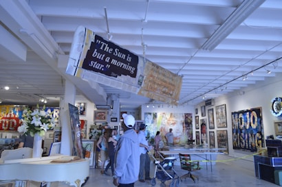 An art gallery with various paintings and artworks displayed on the walls. People are casually wandering around, observing the art. A prominent sign with the quote 'The Sun is but a morning star' hangs from the ceiling. The interior features high white ceilings with beams and an arrangement of flowers on a table.
