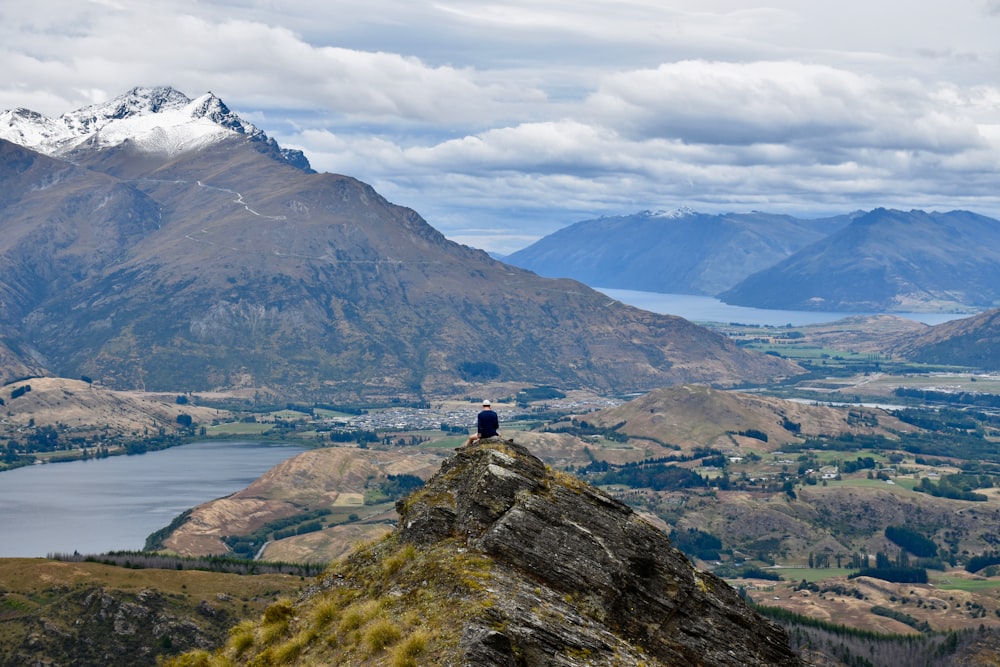 person standing on hill facing mountain and body of water under white and blue sky