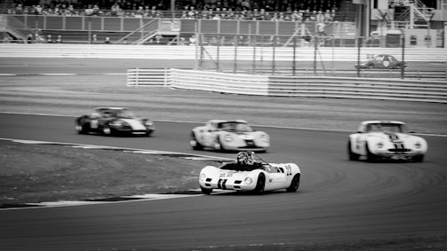 A black and white photograph of classic race cars navigating a racetrack. Several vintage vehicles are captured in motion as they speed around a curve, with stands and spectators in the background.