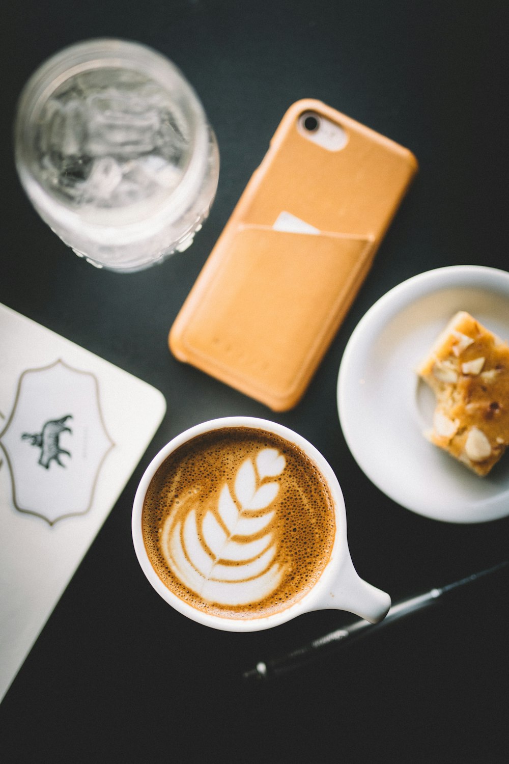 selective focus photo of cup of cappuccino beside smartphone, saucer, and glass