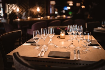 A table set elegantly with multiple wine glasses, cutlery, and dark napkins, surrounded by upholstered chairs. Warm ambient lighting casts a cozy glow, with blurred background suggesting a restaurant or dining setting. A small candle adds an intimate touch.