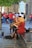 Street musicians are performing with string instruments, including a violin and cello, seated on a bench. They are dressed in casual clothing, with one wearing a wide-brimmed hat. A small crowd gathers around them, with many people wearing red shirts, possibly indicating a group or event.