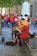 Street musicians are performing with string instruments, including a violin and cello, seated on a bench. They are dressed in casual clothing, with one wearing a wide-brimmed hat. A small crowd gathers around them, with many people wearing red shirts, possibly indicating a group or event.
