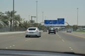A highway scene featuring several cars driving under clear blue road signs that indicate directions to places like Abu Dhabi, Dubai, and Al Ain. Palm trees line both sides of the road, and the sky is bright and clear.