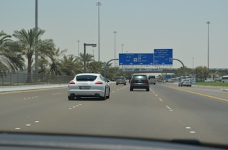A highway scene featuring several cars driving under clear blue road signs that indicate directions to places like Abu Dhabi, Dubai, and Al Ain. Palm trees line both sides of the road, and the sky is bright and clear.