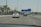 A highway scene featuring several cars driving under clear blue road signs that indicate directions to places like Abu Dhabi, Dubai, and Al Ain. Palm trees line both sides of the road, and the sky is bright and clear.
