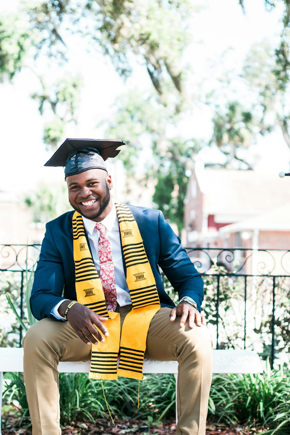 A smiling man in a graduation attire is sitting on a bench in a garden.