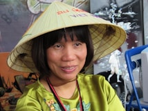 A person wearing a traditional conical hat with colorful writing. The individual has short black hair and is dressed in a vibrant green outfit with embroidered details. The background features a mixture of abstract and cultural art elements along with a blue chair.