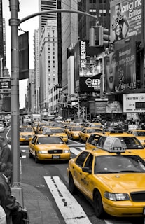 A busy urban scene featuring a fleet of yellow taxis driving through a bustling city street. Skyscrapers line the street, and numerous billboards display advertisements for various products and shows.