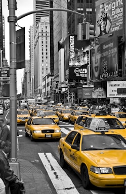 A busy urban scene featuring a fleet of yellow taxis driving through a bustling city street. Skyscrapers line the street, and numerous billboards display advertisements for various products and shows.