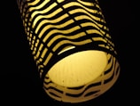 A cylindrical lamp with a warm yellow glow is enclosed in a lampshade with a black wavy pattern. The contrast between the bright light and dark pattern creates a striking visual effect.