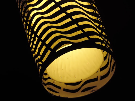 A cylindrical lamp with a warm yellow glow is enclosed in a lampshade with a black wavy pattern. The contrast between the bright light and dark pattern creates a striking visual effect.