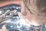 A close-up view of a young child drinking water from a fountain. The focus is on the child's face and the stream of water, with blurred background. The child's hair is slightly wet, indicating playfulness and refreshment.
